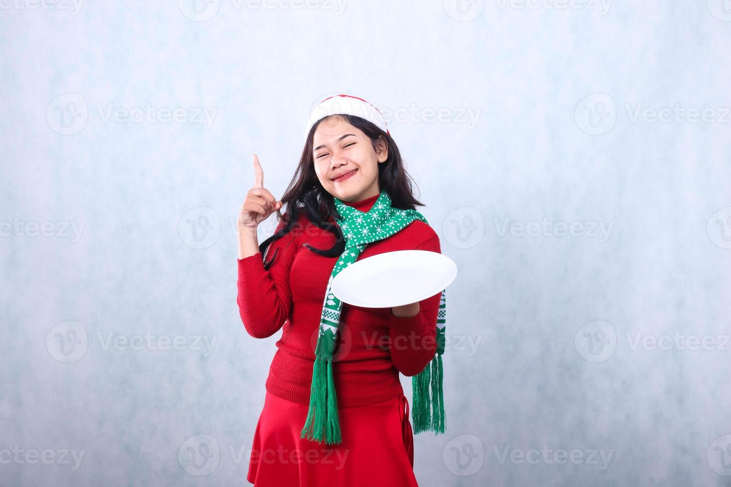 asian girl wearing red sweater with Santa hat and scarf, cheerful looking at camera hand holding white plate and pointing up, celebrating merry christmas and new year isolated on white background photo