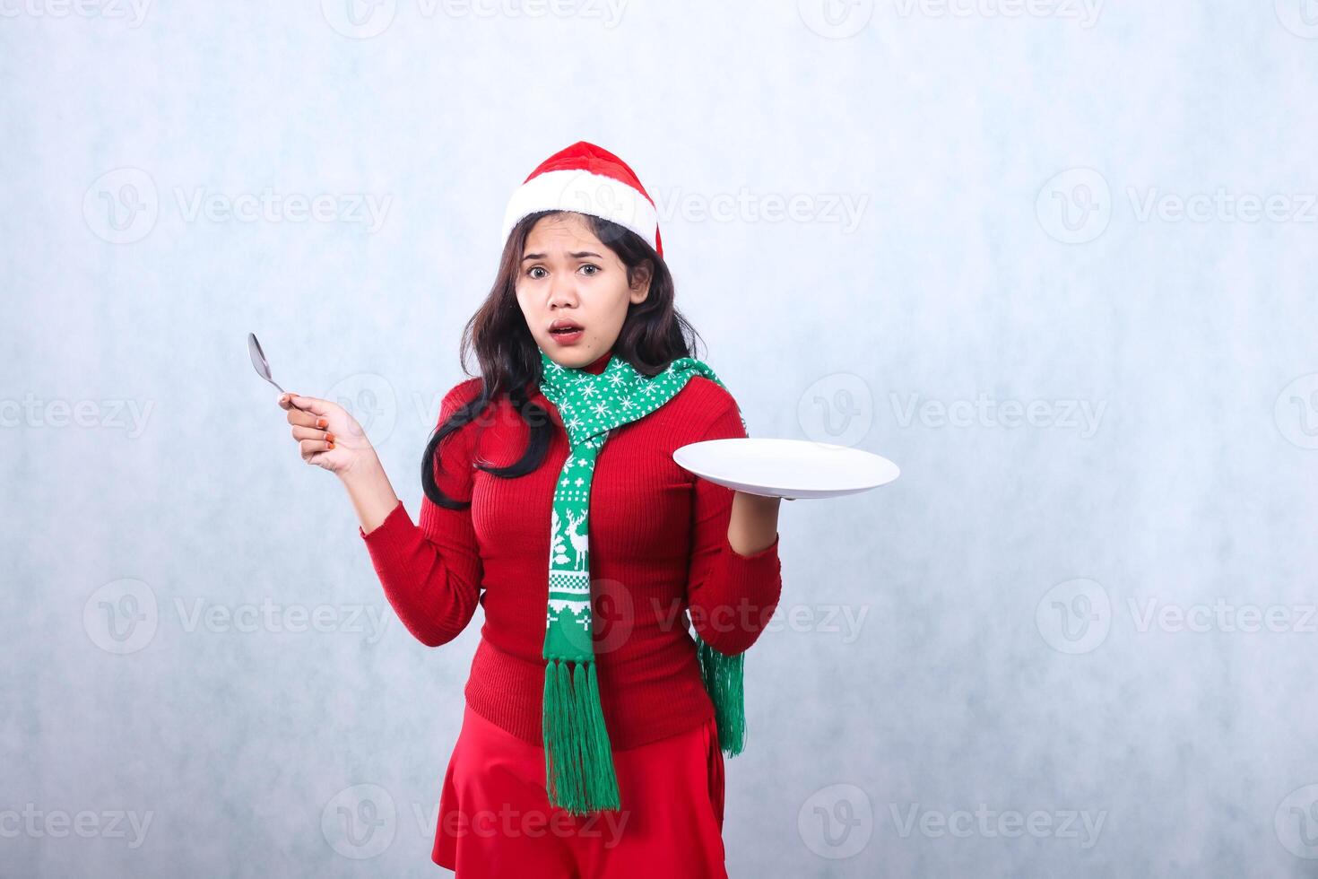 gesture adult woman wearing red sweater with Santa hat and scarf, surprised to camera carrying white plate and holding spoon to the side, celebrating merry christmas isolated on white background photo