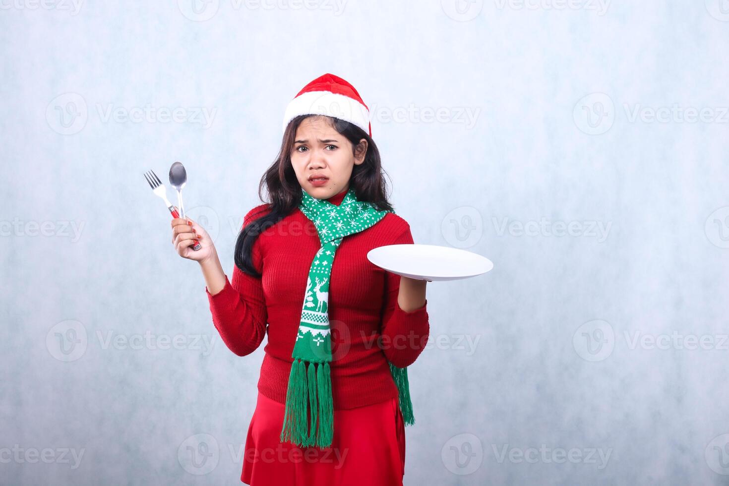 gesture indonesia girl wearing red sweater with Santa hat and scarf, scared and cheerful to camera carry white plate and hold spoon fork, celebrate merry christmas isolated on white background photo
