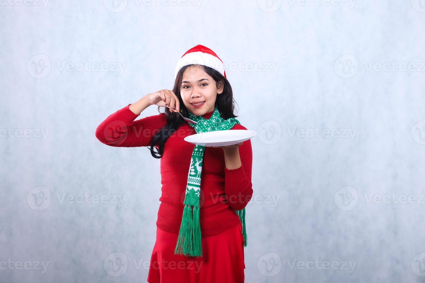 gesture asian woman wearing red sweater with Santa hat and scarf, smile to camera carrying white plate and scooping food with spoon, celebrating merry christmas isolated on white background photo