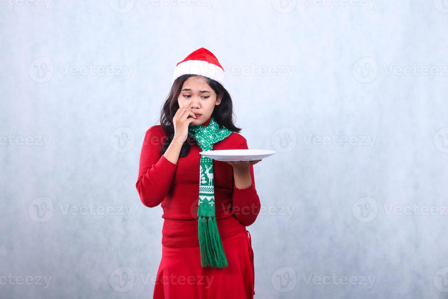 portrait woman wearing red sweater with Santa hat and scarf, scared to the plate holding white plate while sticking mouth, celebrating merry christmas and new year isolated on white background photo