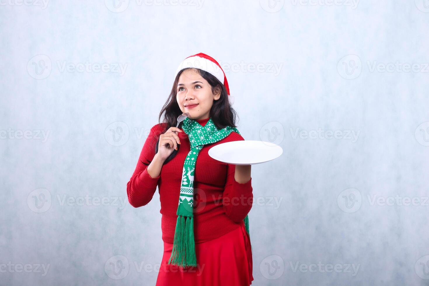 gesture young woman wearing red sweater with Santa hat and scarf, smiling while thinking carry white plate and hold spoon, celebrating merry christmas and happy new year isolated on white background photo
