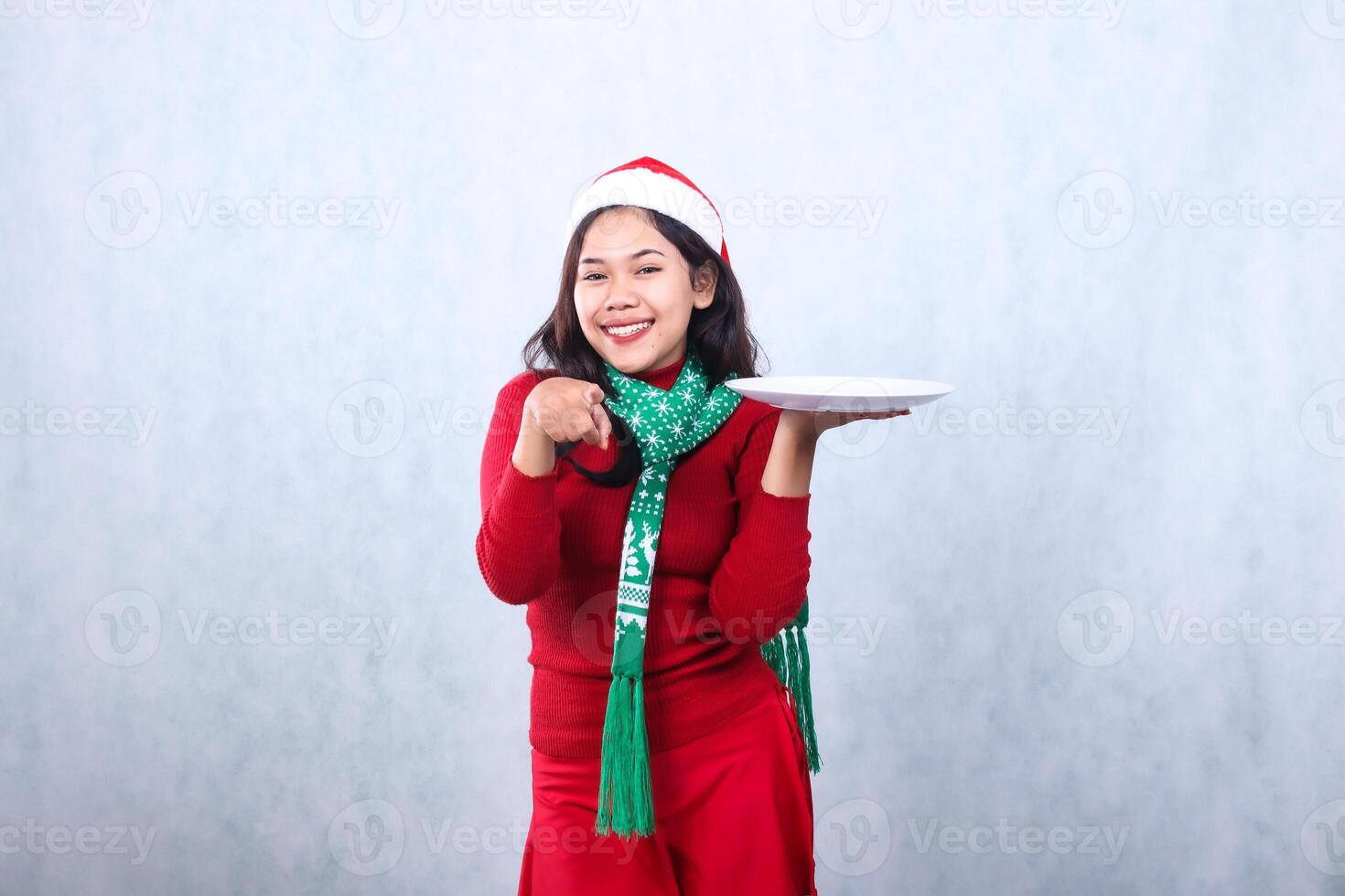 female wearing red sweater with Santa hat and scarf, cheerful looking at camera, hand lifting white plate and pointing forward, celebrating merry christmas and new year isolated on white background photo