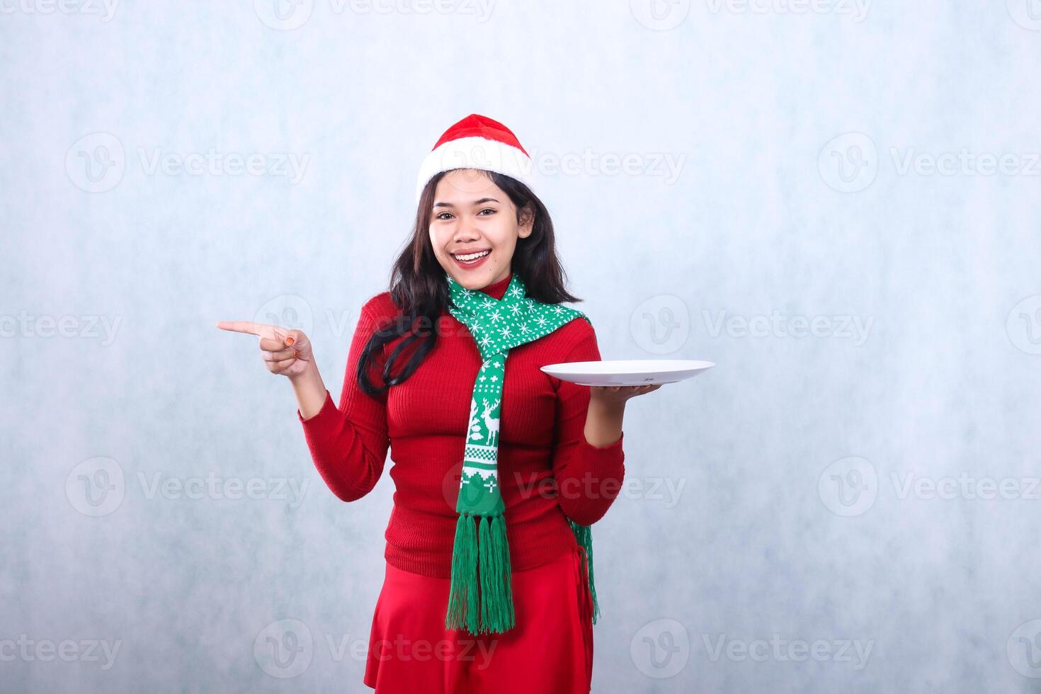 portrait of Asian girl wearing red sweater red sweater with Santa hat and scarf celebrating merry christmas, cheerful to camera holding white plate while pointing right, isolated on white background photo