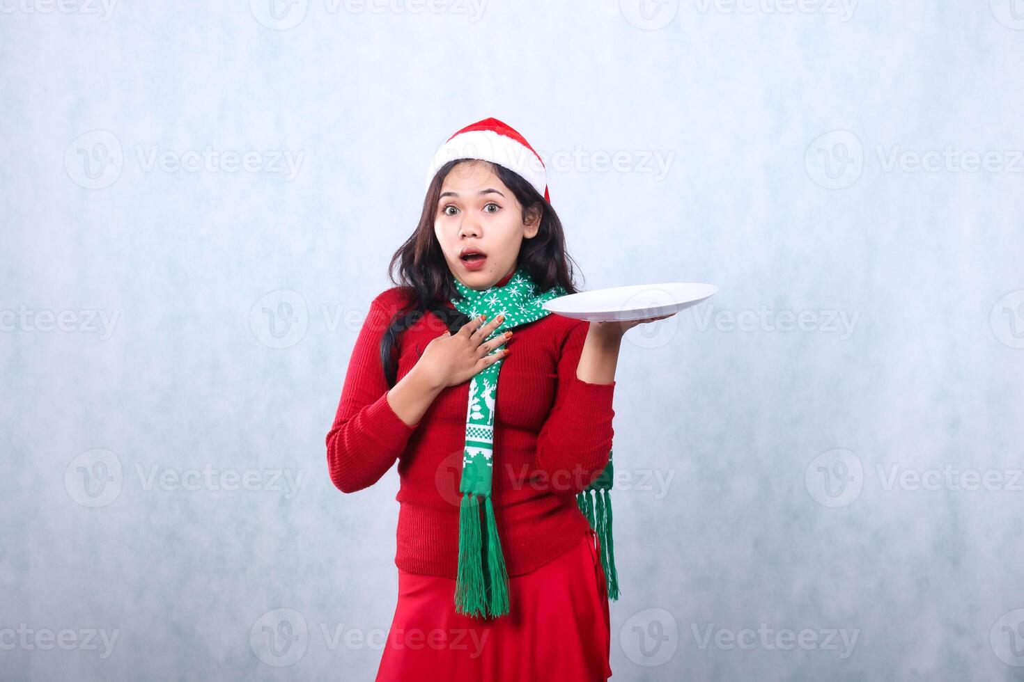 woman expression wearing red sweater red sweater with Santa hat and scarf celebrating merry christmas, shocked looking at camera hand lifting white plate and holding chest, isolated white background photo