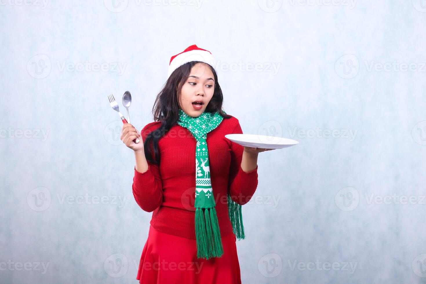 portrait of Asian woman wearing red sweater red sweater with Santa hat and scarf celebrating merry christmas, surprised to food carry white plate and hold spoon fork, isolated on white background photo