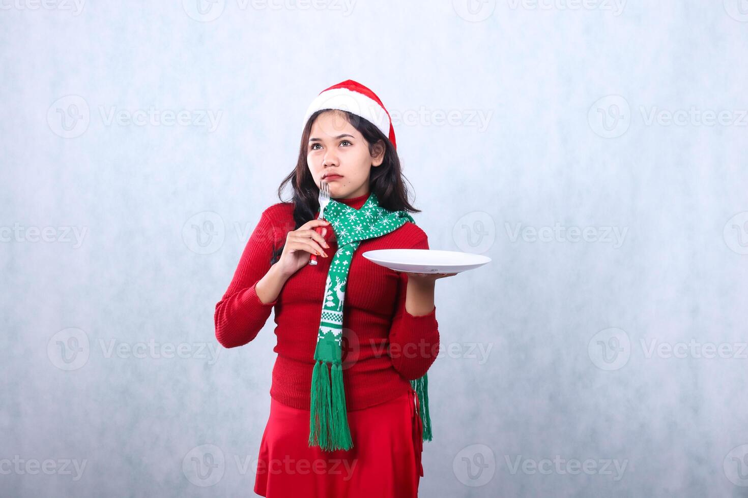 portrait of asian girl wearing red sweater red sweater with santa hat and scarf celebrating merry christmas and new year, looking for ideas carry white plate and hold fork, isolated white background photo