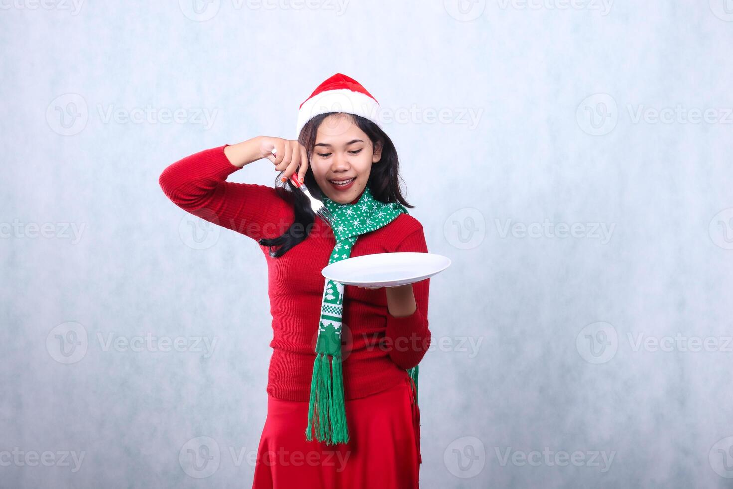 gesture young asian woman wearing red sweater with Santa hat and scarf, cheerful to plate carry white plate and hold fork, celebrating merry christmas and new year isolated on white background photo