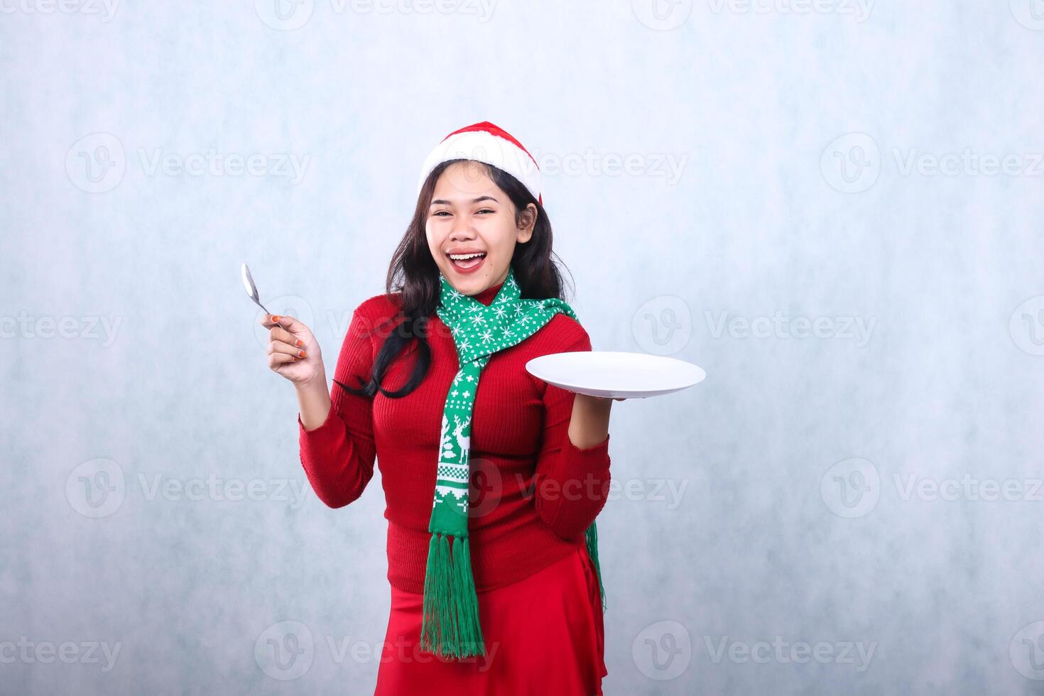 Charming indonesia female wearing red sweater with Santa hat and scarf, happy to camera carrying white plate and holding spoon, celebrating merry christmas and new year isolated on white background photo