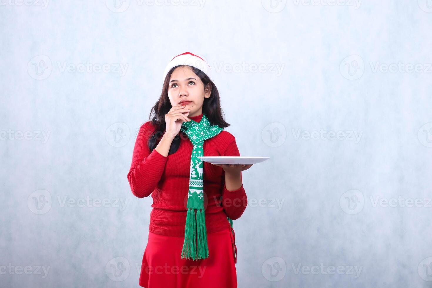 portrait of Asian girl wearing red sweater red sweater with Santa hat and scarf celebrating merry christmas, thinking about something while holding white plate, isolated on white background photo