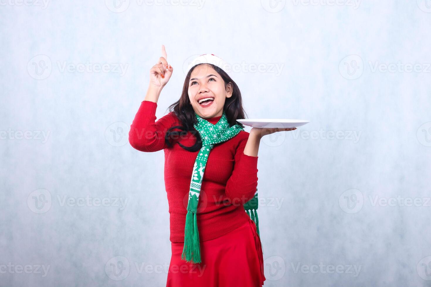 expression of asian woman wearing red sweater red sweater with Santa hat and scarf celebrating merry christmas, cheerful look up hand lift white plate and point up, isolated on white background photo