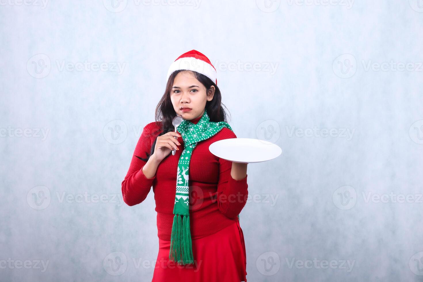 Charming young woman wearing red sweater with Santa hat and scarf, looking at camera, carrying white plate and holding spoon, celebrating merry christmas and new year isolated on white background photo