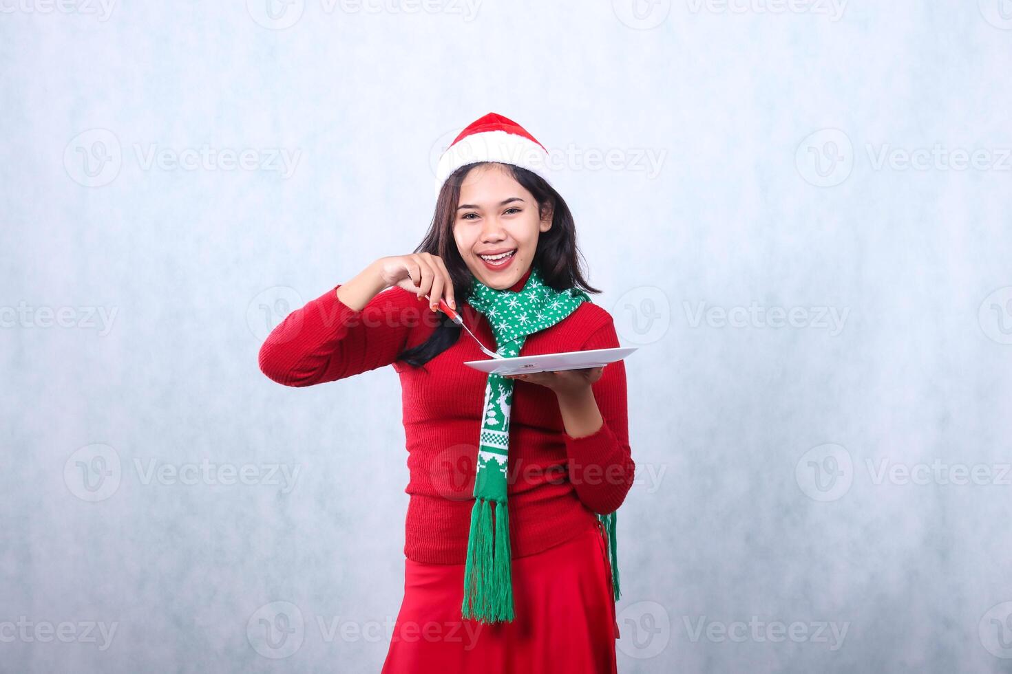 gesture young asian woman wearing red sweater with Santa hat and scarf, cheerful to camera carrying white plate and scooping food with fork, celebrating merry christmas isolated on white background photo