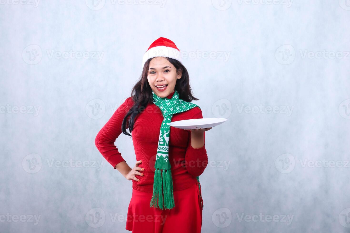 gesture young asian woman wearing red sweater with Santa hat and scarf, cheerful to camera holding white plate and waist, celebrating merry christmas and new year isolated on white background photo