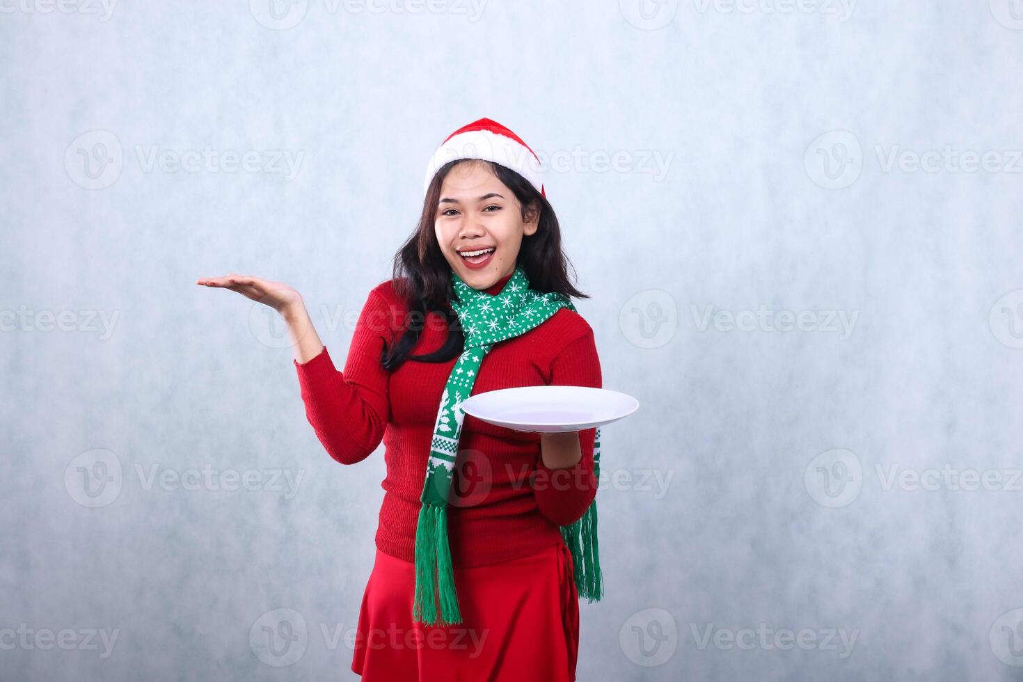 gesture woman wearing red sweater with Santa hat and scarf, cheerful looking at camera hand holding white plate and palm to right, celebrating merry christmas and new year isolated on white background photo