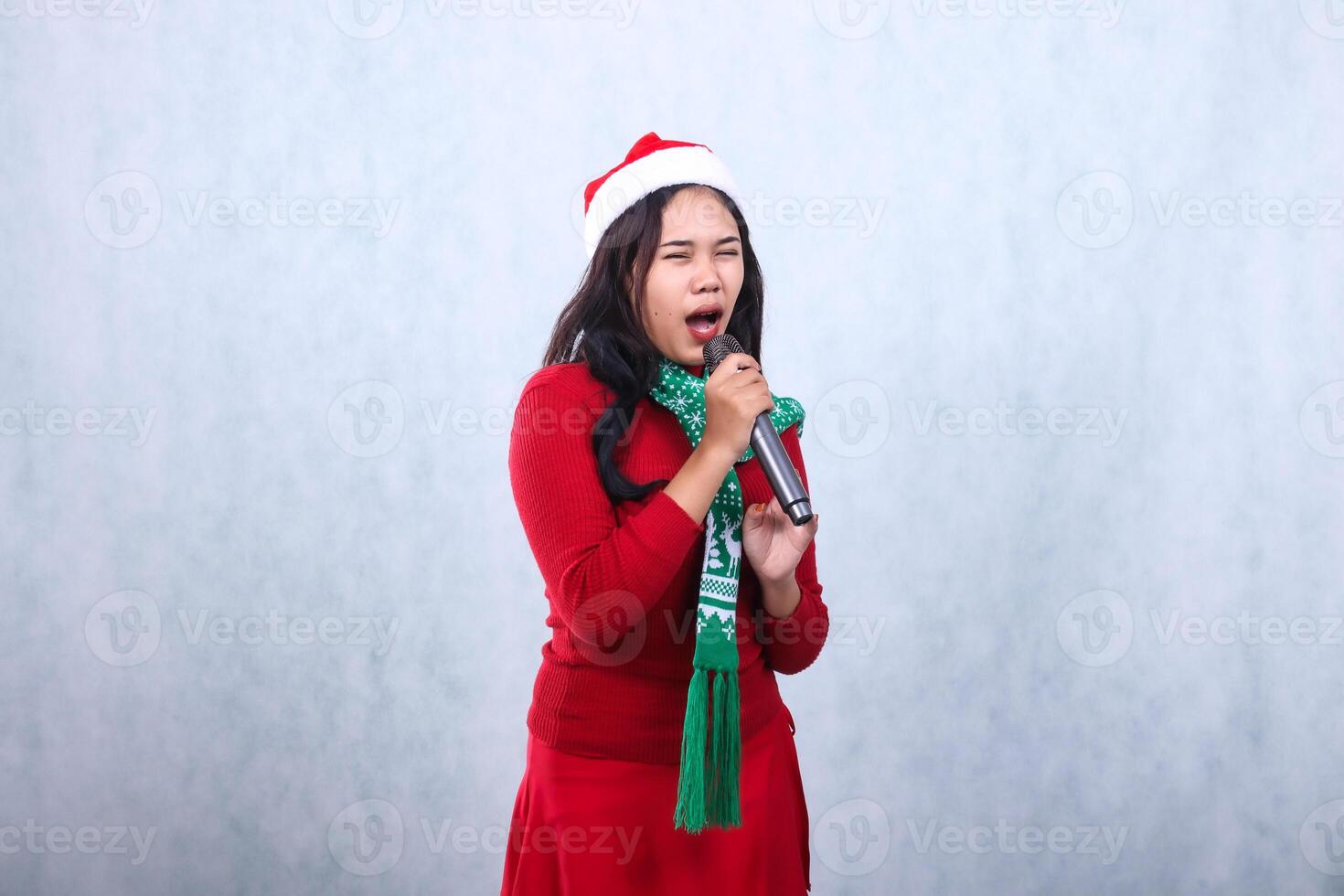 portrait of Asian female wearing red sweater with Santa hat and scarf celebrating merry christmas, shouting singing closing eyes both hands holding karaoke mic, isolated on white background photo