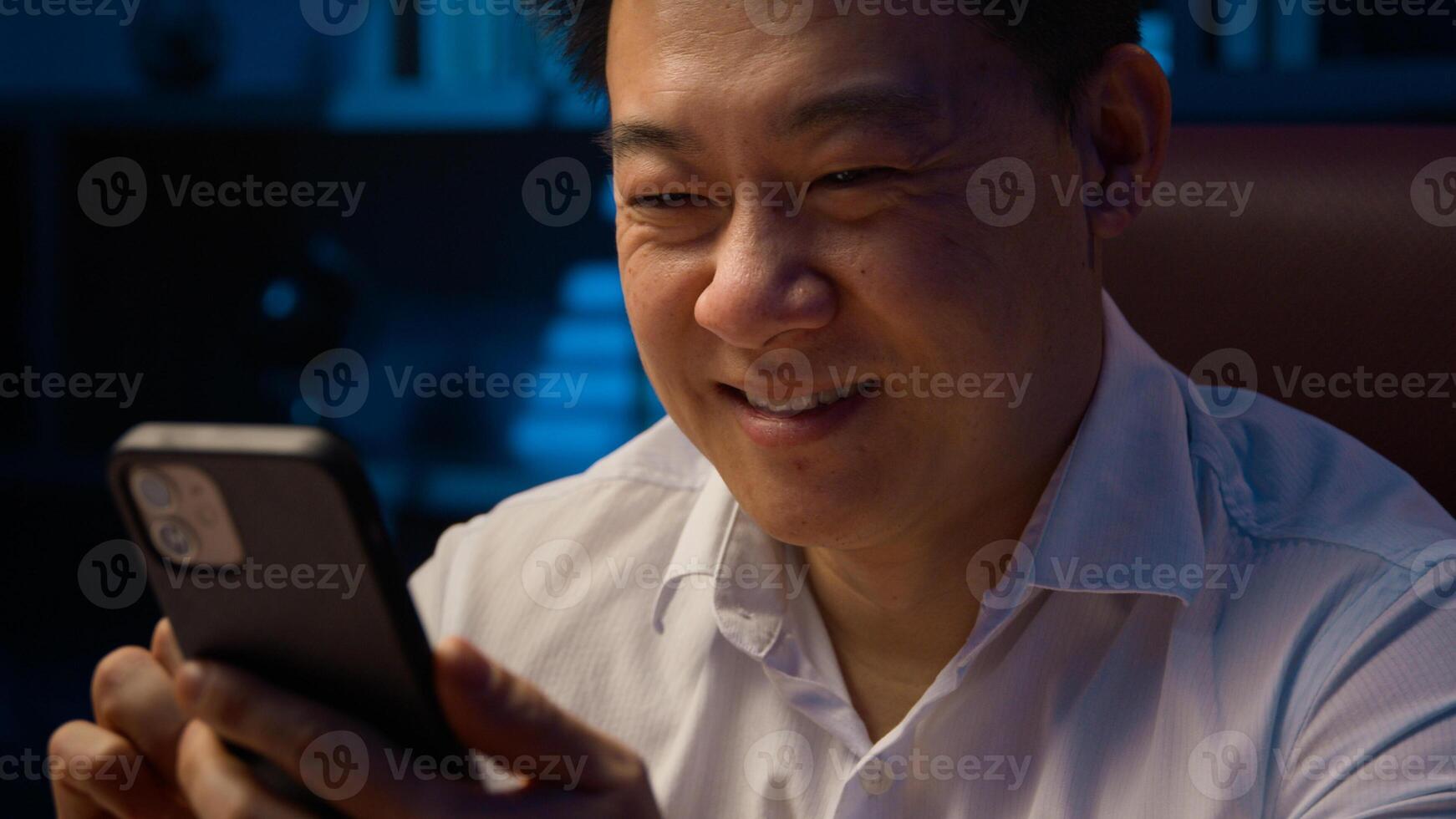 Happy smiling Asian middle-aged business man Korean adult businessman Chinese worker Japanese employer at night evening home office browsing mobile phone scrolling smartphone chatting cellphone app photo