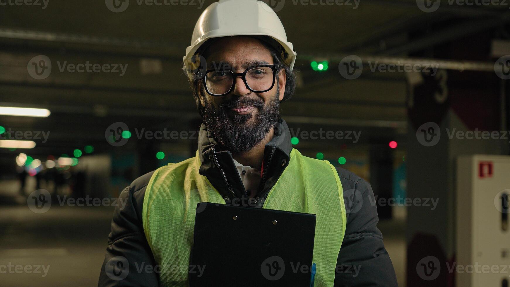 Indian contractor mechanic guy in helmet and glasses smile posing with clipboard folder. Arabian bearded man in eyeglasses worker male specialist manufacture work. Engineer in hard hat safety uniform photo
