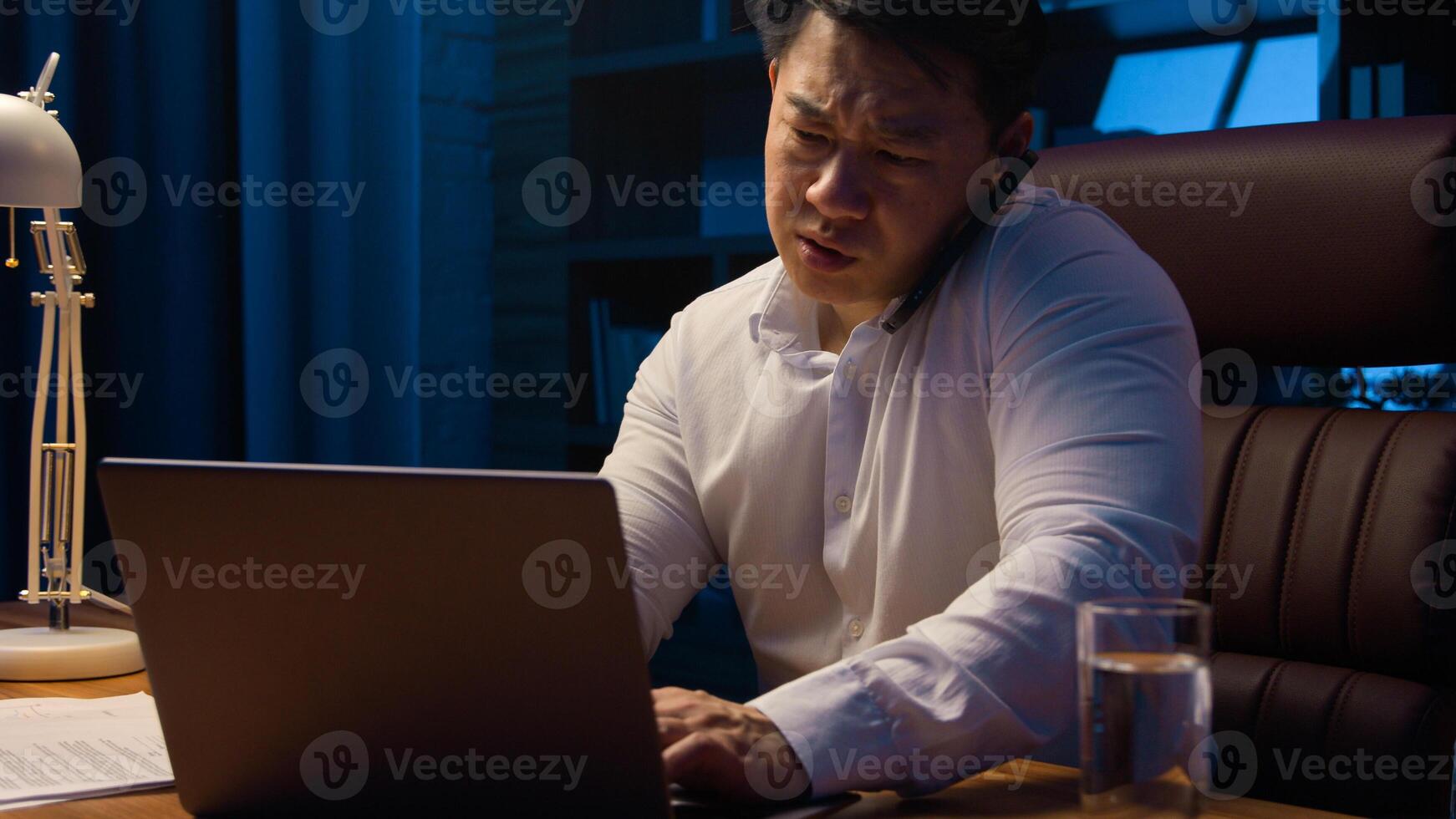 Tired angry Asian business man talking mobile phone at night evening office check documents working laptop overworked stressed Korean businessman executive manager arguing with call computer paperwork photo