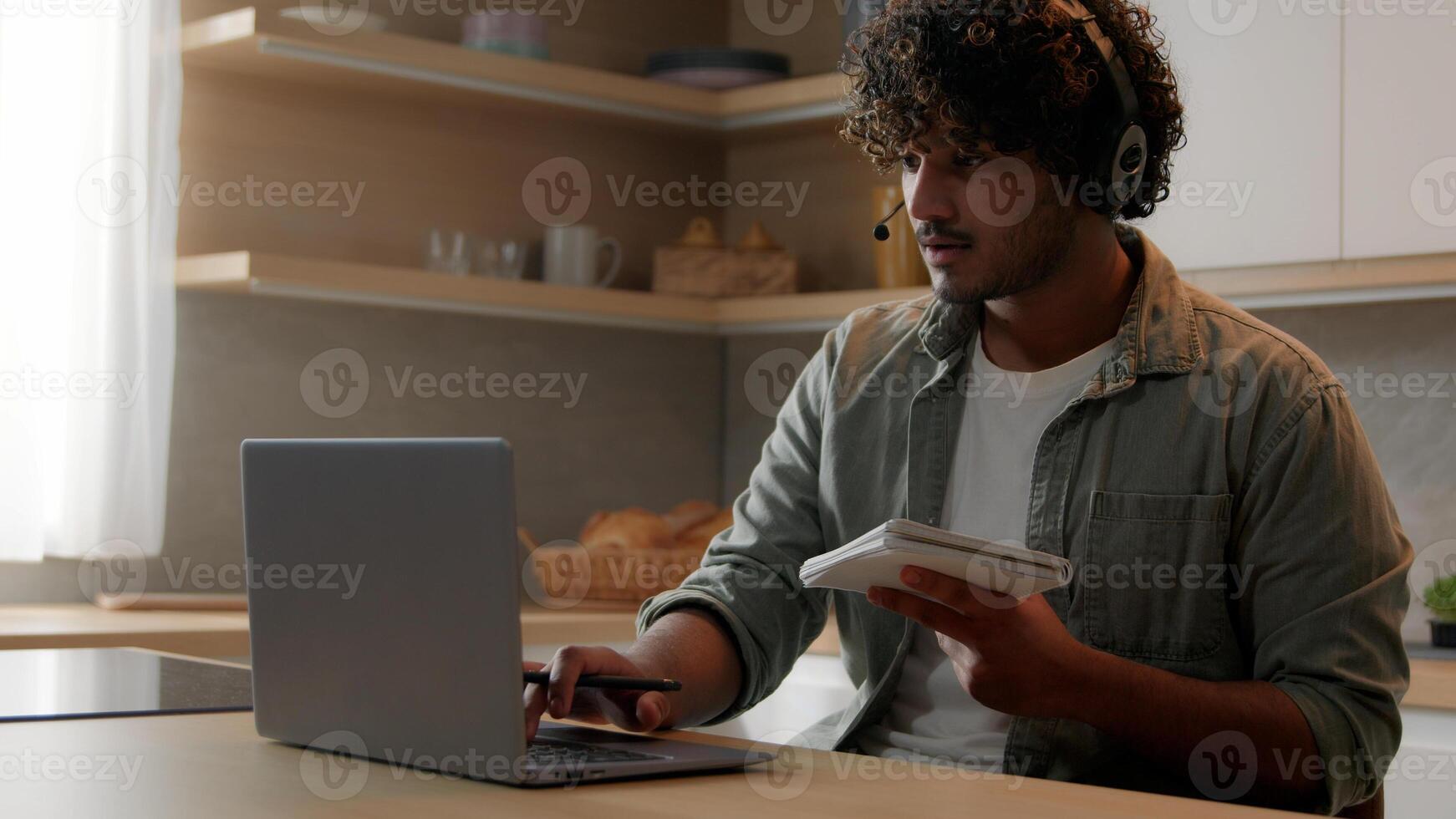 Indian man in headset look app pc browsing typing laptop write ideas questions notes review research sit at home kitchen. Arabian guy learn language improve knowledge self-education distant e-learning photo