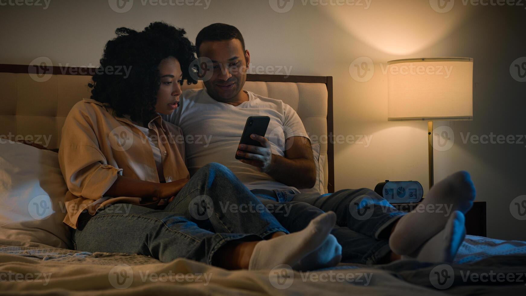 African American couple man and woman husband wife boyfriend girlfriend using mobile phone together lying in bed at night enjoying relaxing scrolling smartphone technology watching social media photo