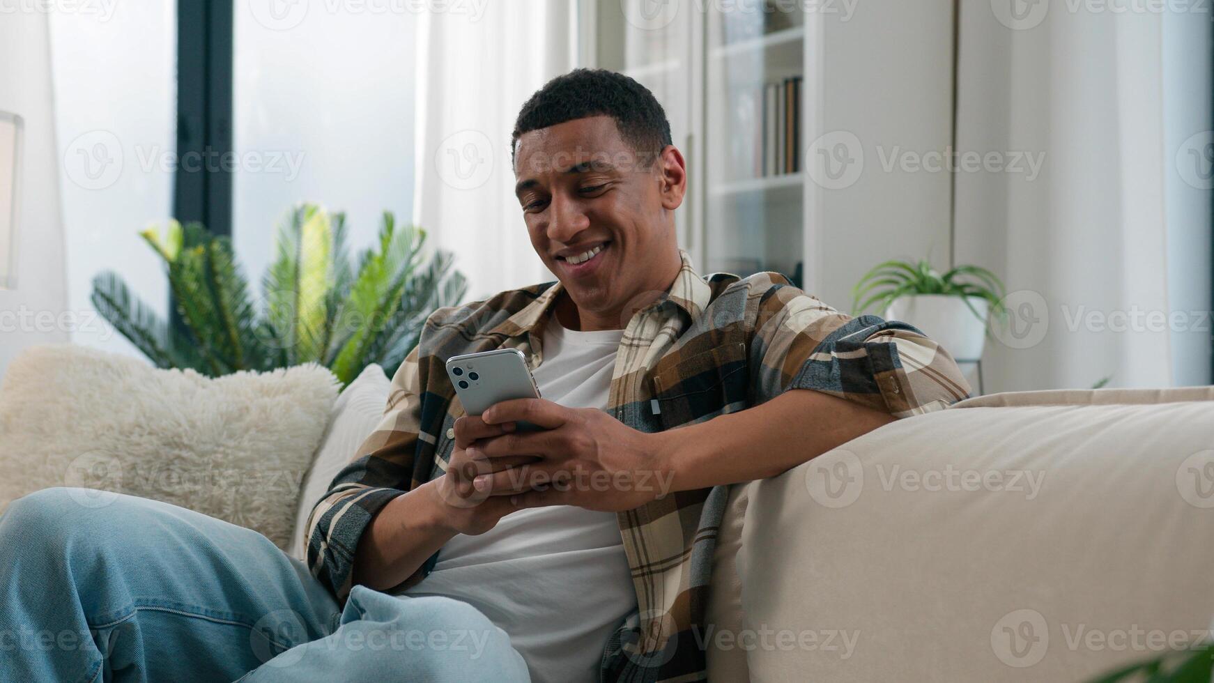 African American guy ethnic happy smiling man relaxing on comfortable couch sofa at home using mobile application scrolling phone app chatting social network online web surfing internet smartphone net photo
