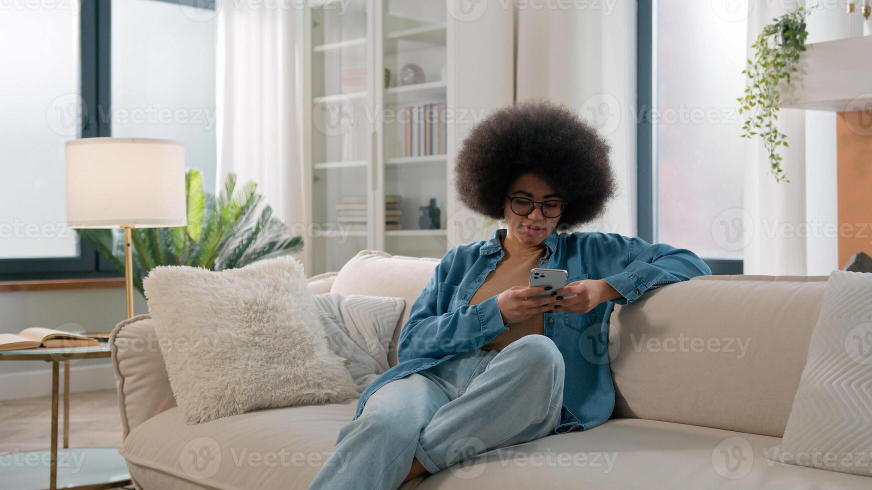 Relaxed calm African American ethnic woman typing swipe touchscreen scrolling mobile phone check social media cellphone app girl choosing purchase shopping in online e-commerce application at home photo