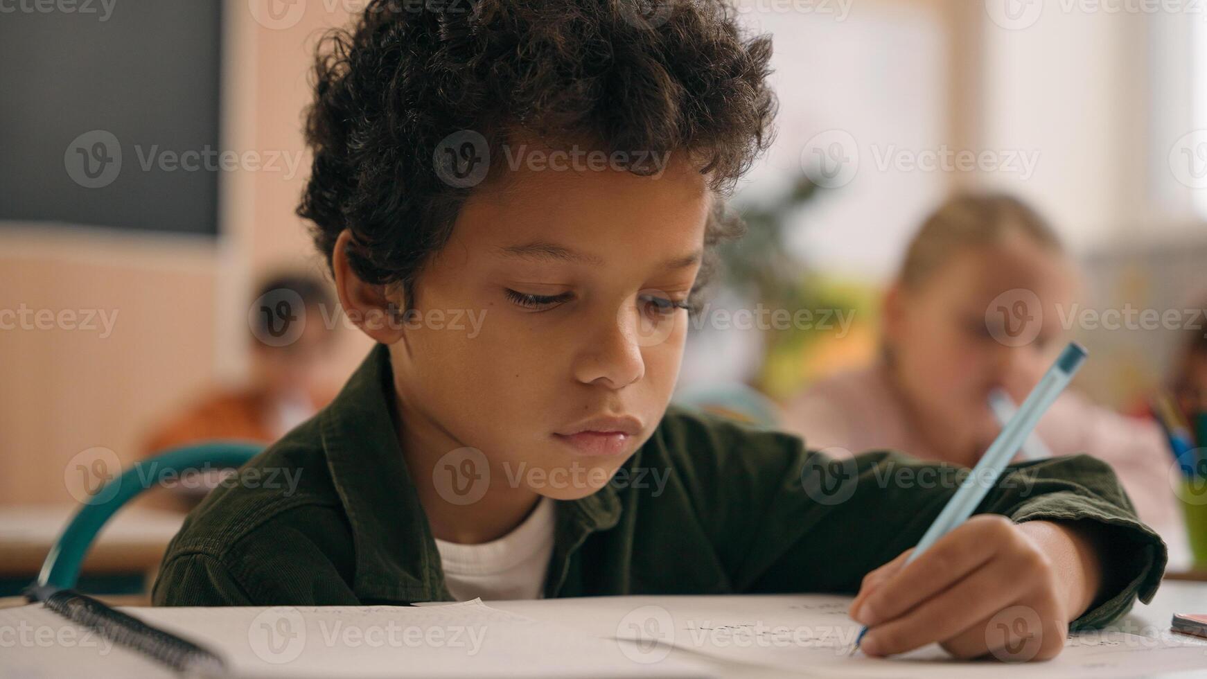 Little African American guy boy writing notes task class lesson primary school write in copybook kid schoolboy son child pupil student learning listen teacher studying education sitting at table desk photo