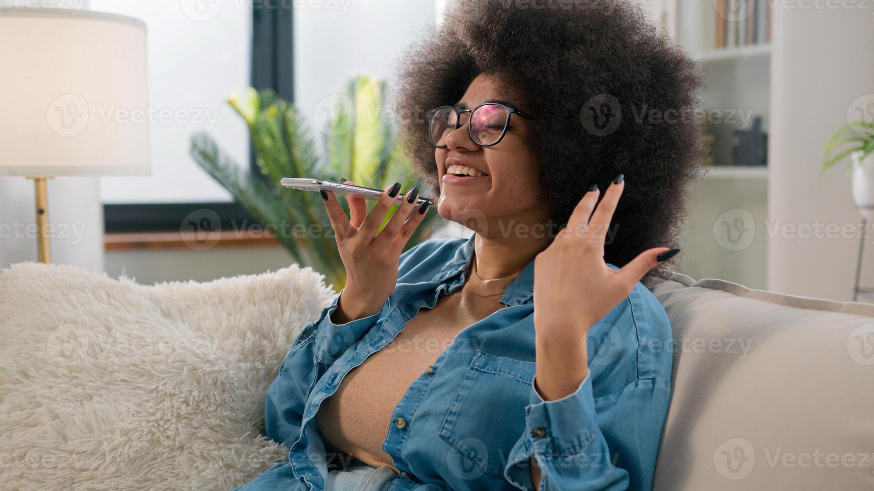 Ethnic girl African American woman talk virtual assistant on mobile phone smiling lady recording answering audio message voice mail in social network for friend using loudspeaker talking at home sofa photo