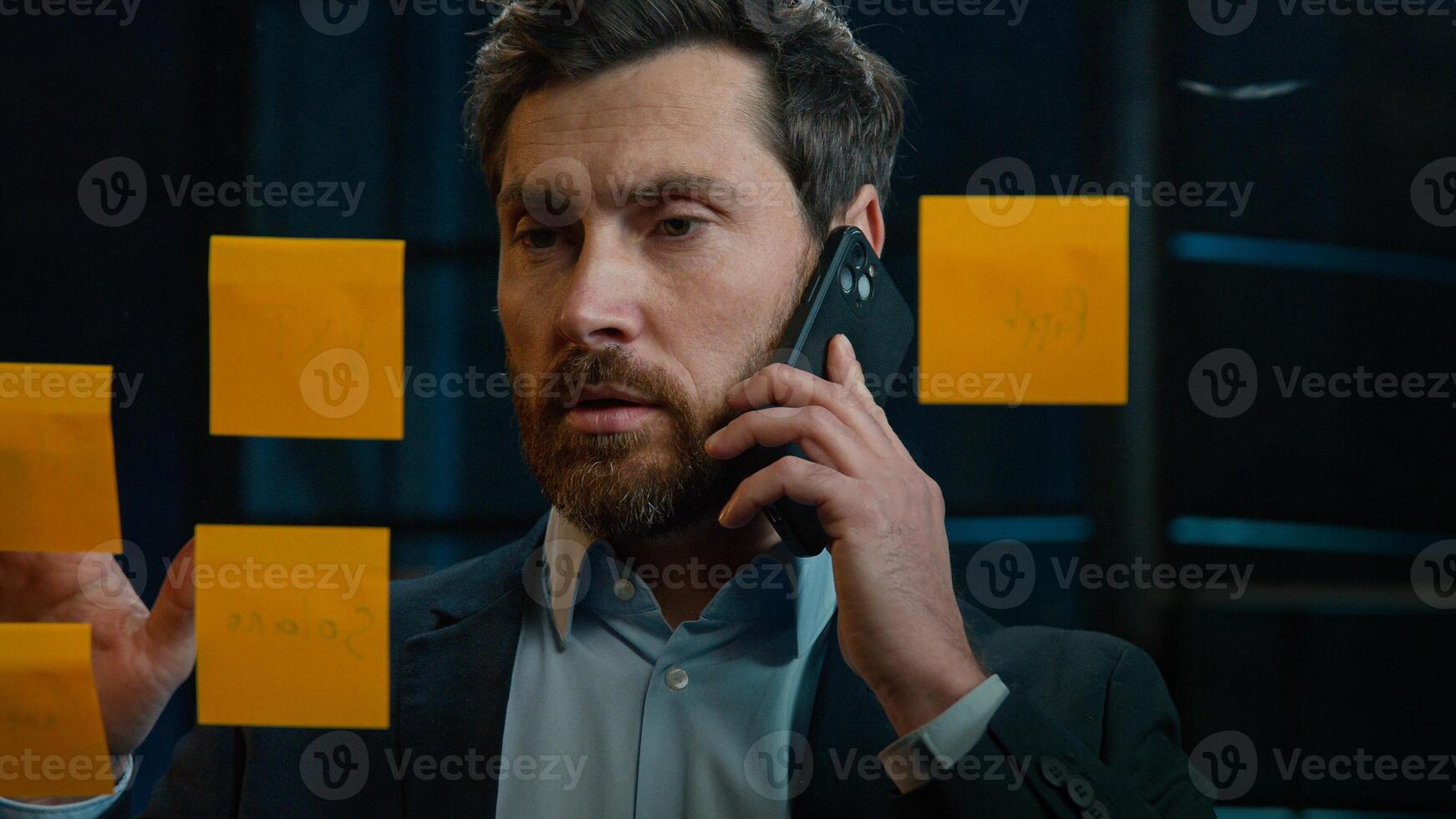 Caucasian businessman marketing assistant middle-aged man multitask talk mobile phone discuss project at office read sticky notes on glass manage business tasks goals analyzing sales strategy research photo