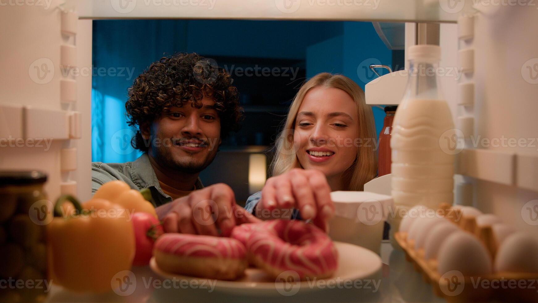 Point of view POV from inside refrigerator multiracial couple diverse Indian guy man and Caucasian girl woman at night kitchen open fridge eating donut unhealthy nutrition diet failure obesity food photo