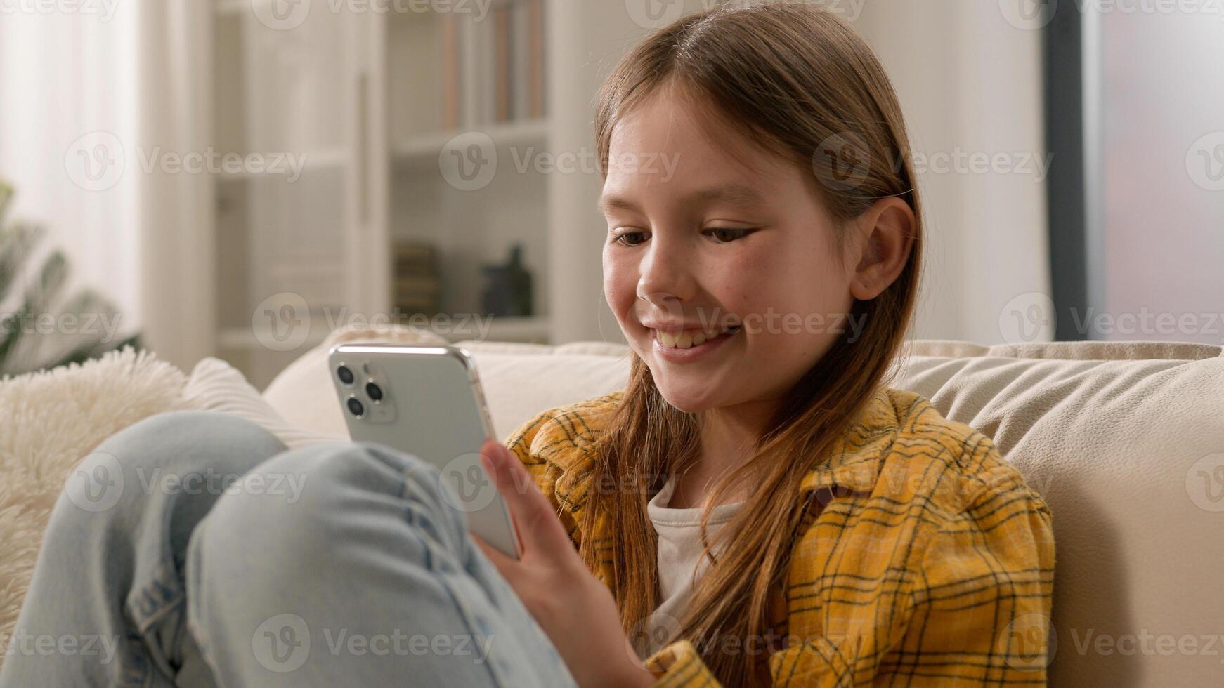 Smiling happy kid girl sending message checking email using mobile phone cute schoolgirl child chatting with friends on social network internet playing online game watching on smartphone at home photo