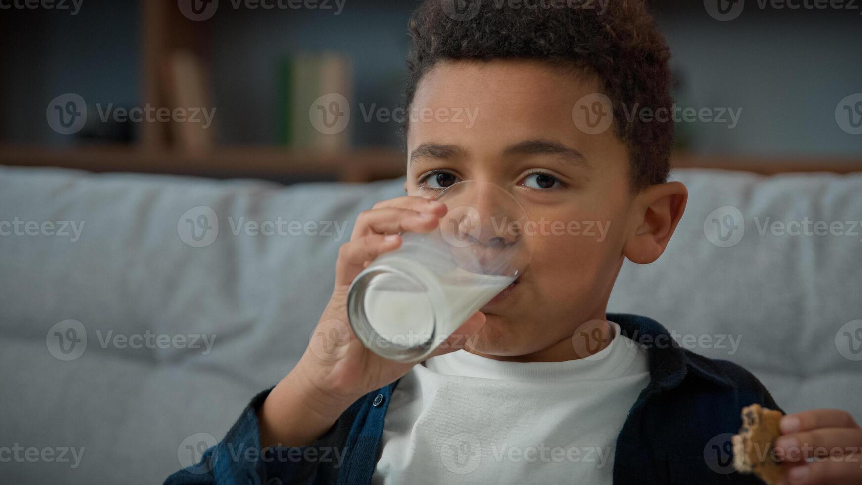 Close up portrait little funny cute kid boy ethnic child drink milk with cookies at home looking to camera. African American son drinking glass of sweet organic yogurt eating biscuit breakfast eat photo