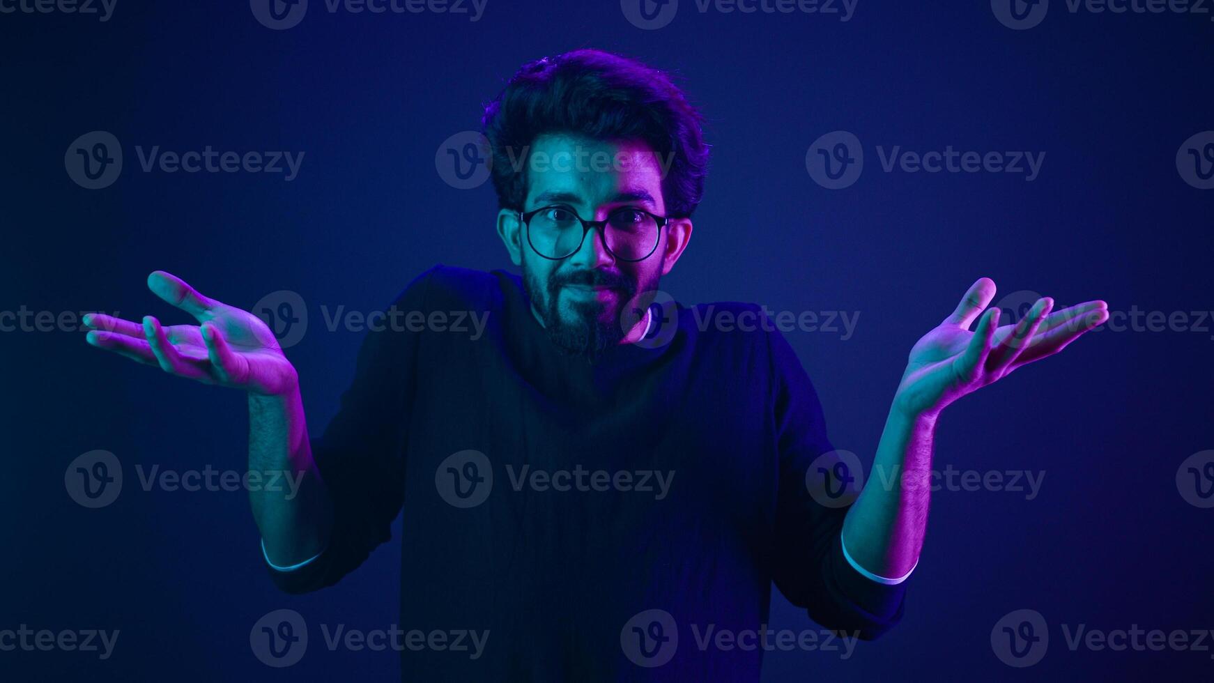 Male portrait in blue neon ultraviolet background Indian man shrug shoulders don't know gesture Arabian male guy confused unsure doubtful shrugging doubt uncertain guess puzzled bemused no clue sign photo