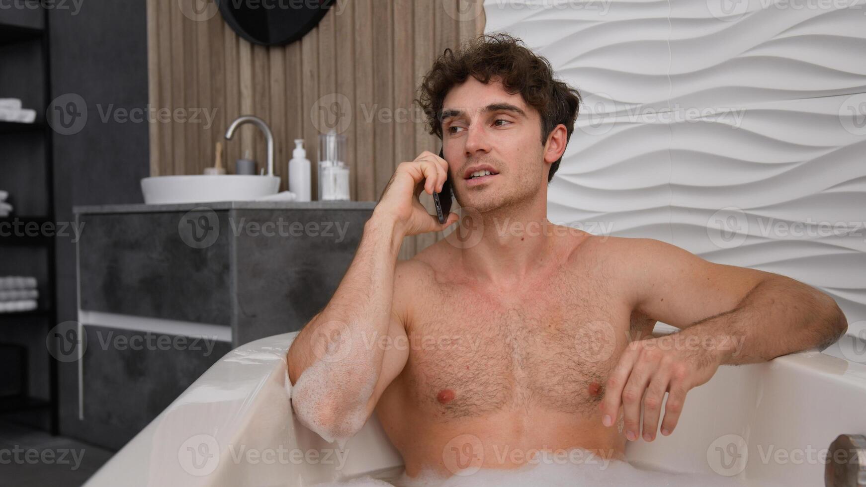 Caucasian man naked male guy relaxing in bath with soapy foam relax rest in bathroom in bathtub talking smartphone call chat discuss talk mobile phone calling speak washing in tub hygiene holiday spa photo