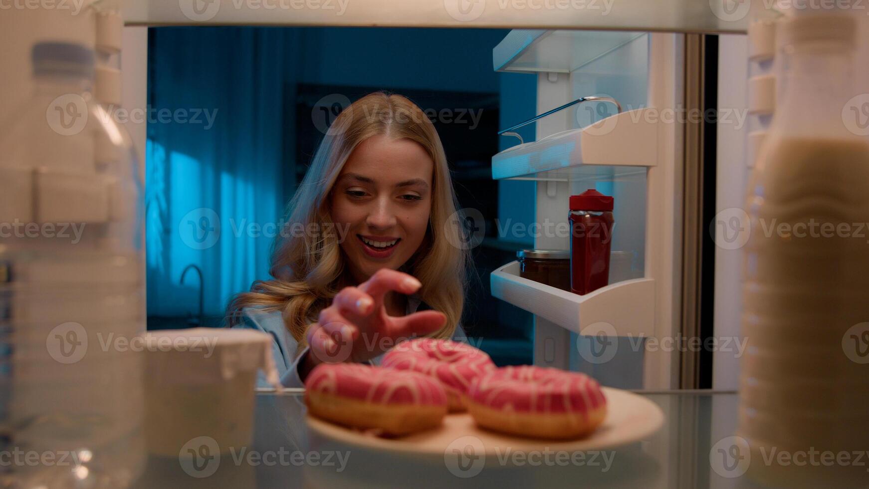 Point of view POV from inside refrigerator hungry Caucasian woman girl at night evening kitchen open fridge take out eating sweet donut unhealthy nutrition obesity diet failure overeating food addict photo