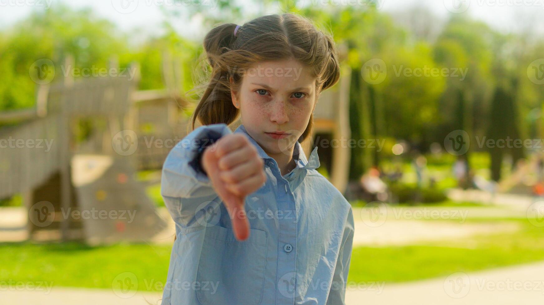 Caucasian girl female child kid little daughter serious sad dissatisfied schoolgirl school pupil teen outdoors park city showing thumb down gesture disapprove not dislike bad negative bullying problem photo