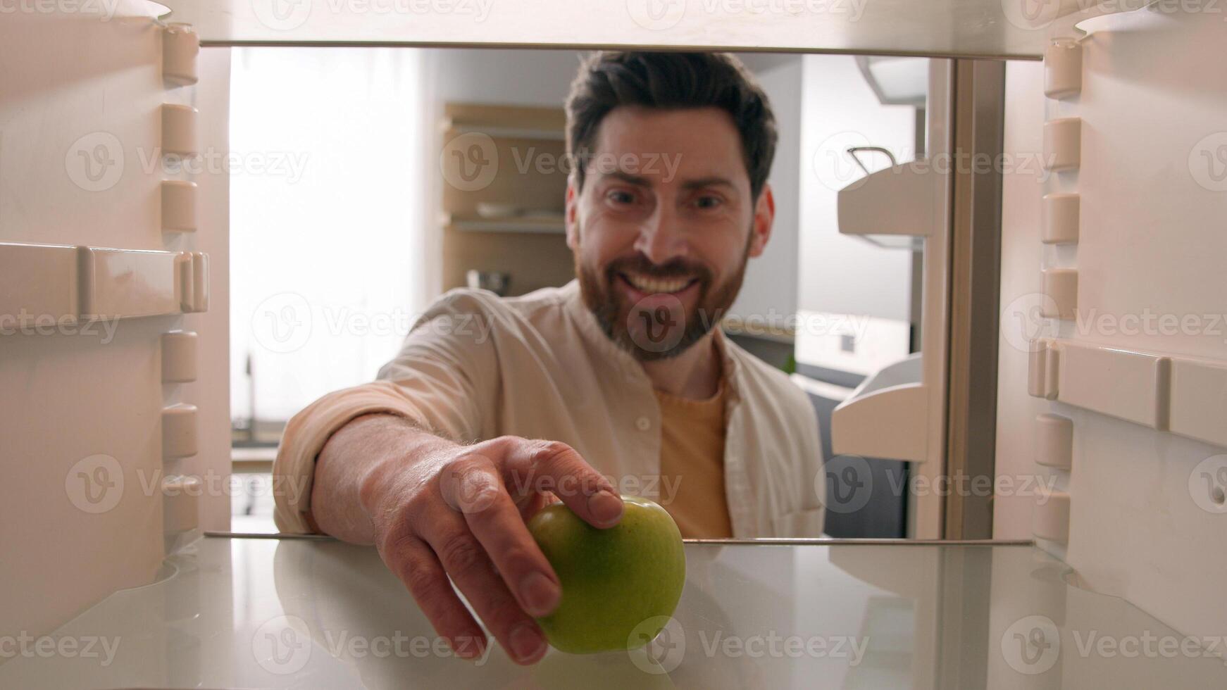 POV point of view from inside refrigerator Caucasian adult happy man at kitchen open empty fridge take last green apple bite eat fresh healthy fruit hungry guy male eating diet food delivery service photo