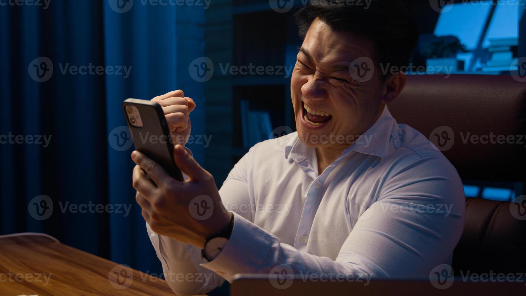 Excited shocked surprised Asian man achievement mobile phone prize offer opportunity happy Korean winner businessman victory smartphone win screaming champion business winning at night evening office photo