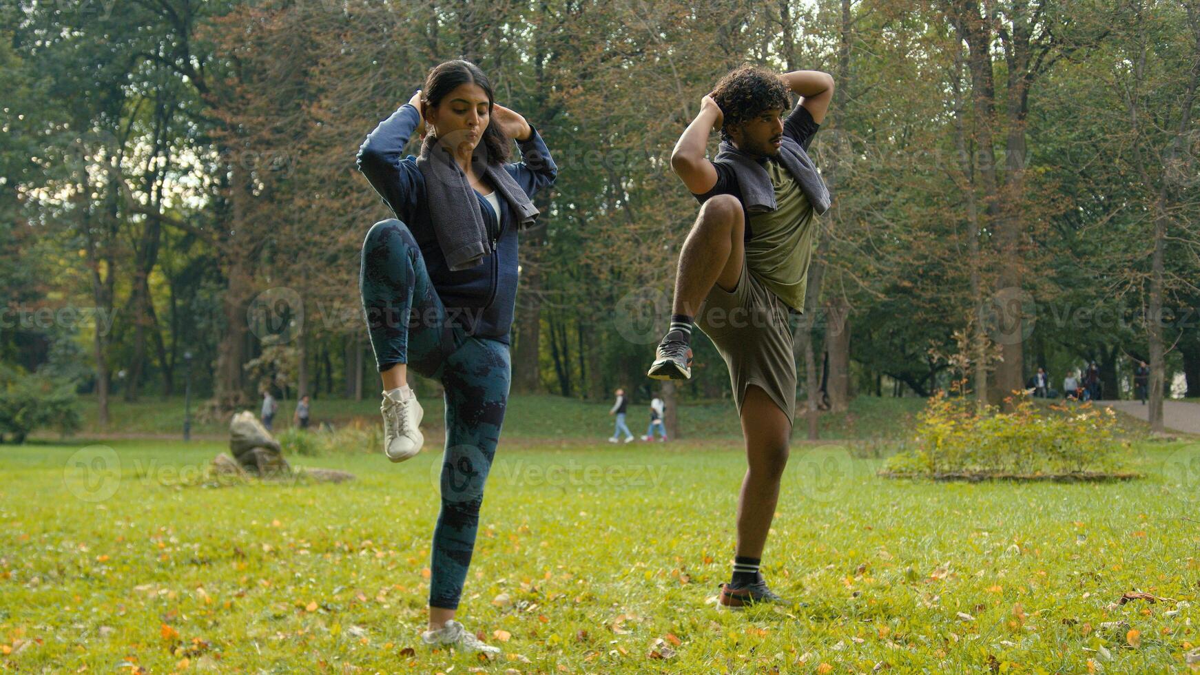 Indian man sportsman athlete instructor personal fitness trainer guy Arabian woman girl sportswoman fit couple sport exercise workout together in park outdoors raise knees to elbows while standing photo