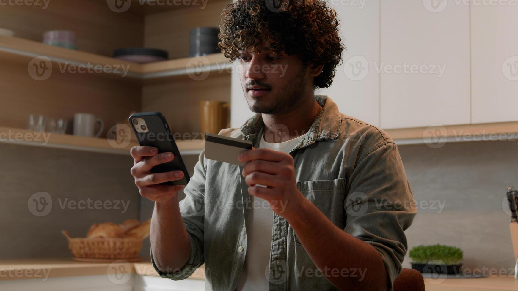 Indian male customer man Arabian guy homeowner sitting at kitchen order food products with smart phone app using bank credit card making purchases at web grocery store delivery service shopping online photo