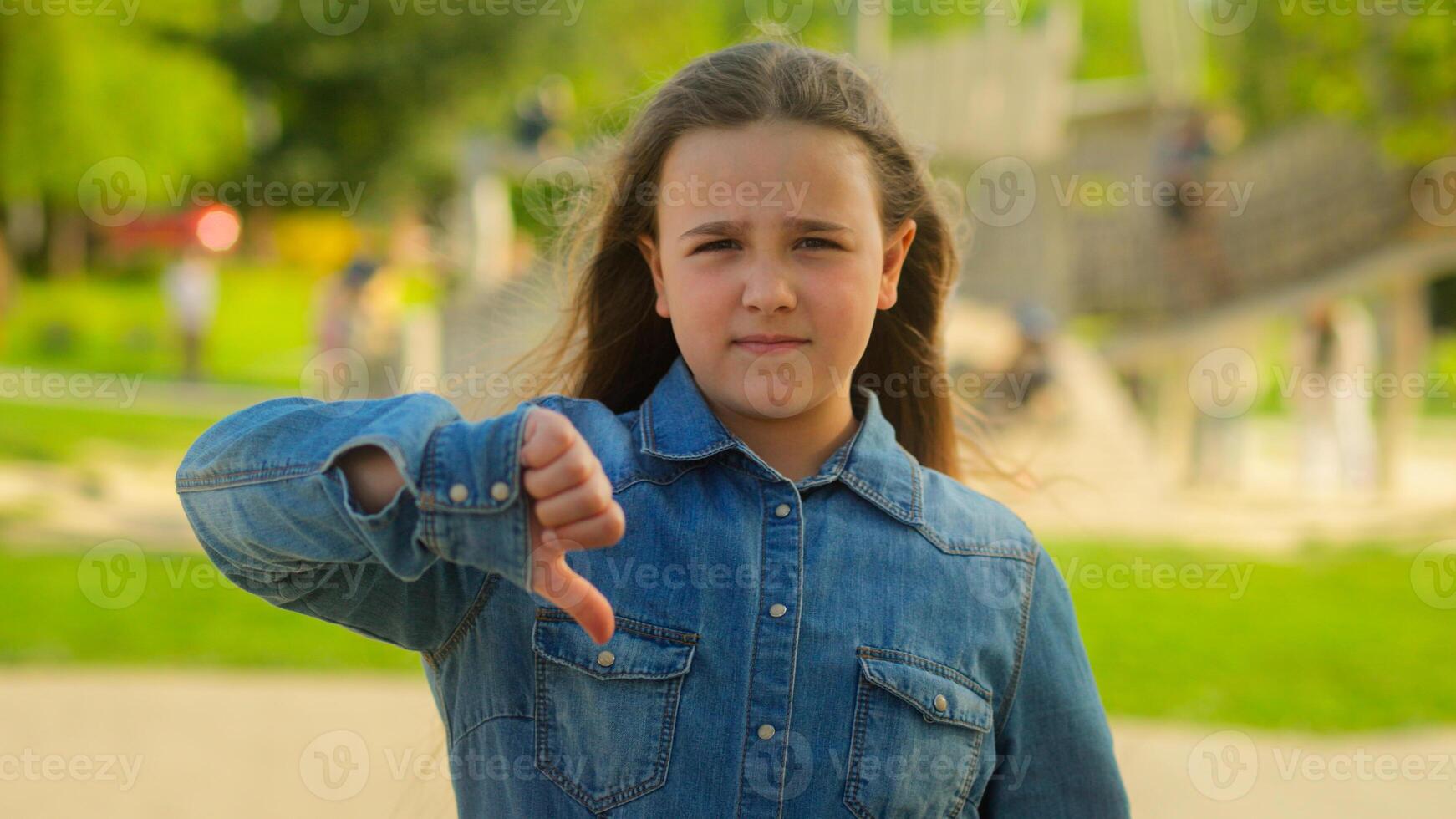 Sad upset displeased little girl schoolgirl child serious kid daughter in city outdoors looking camera showing thumb down finger gesture dislike not low mark disapprove sign childhood custody problem photo