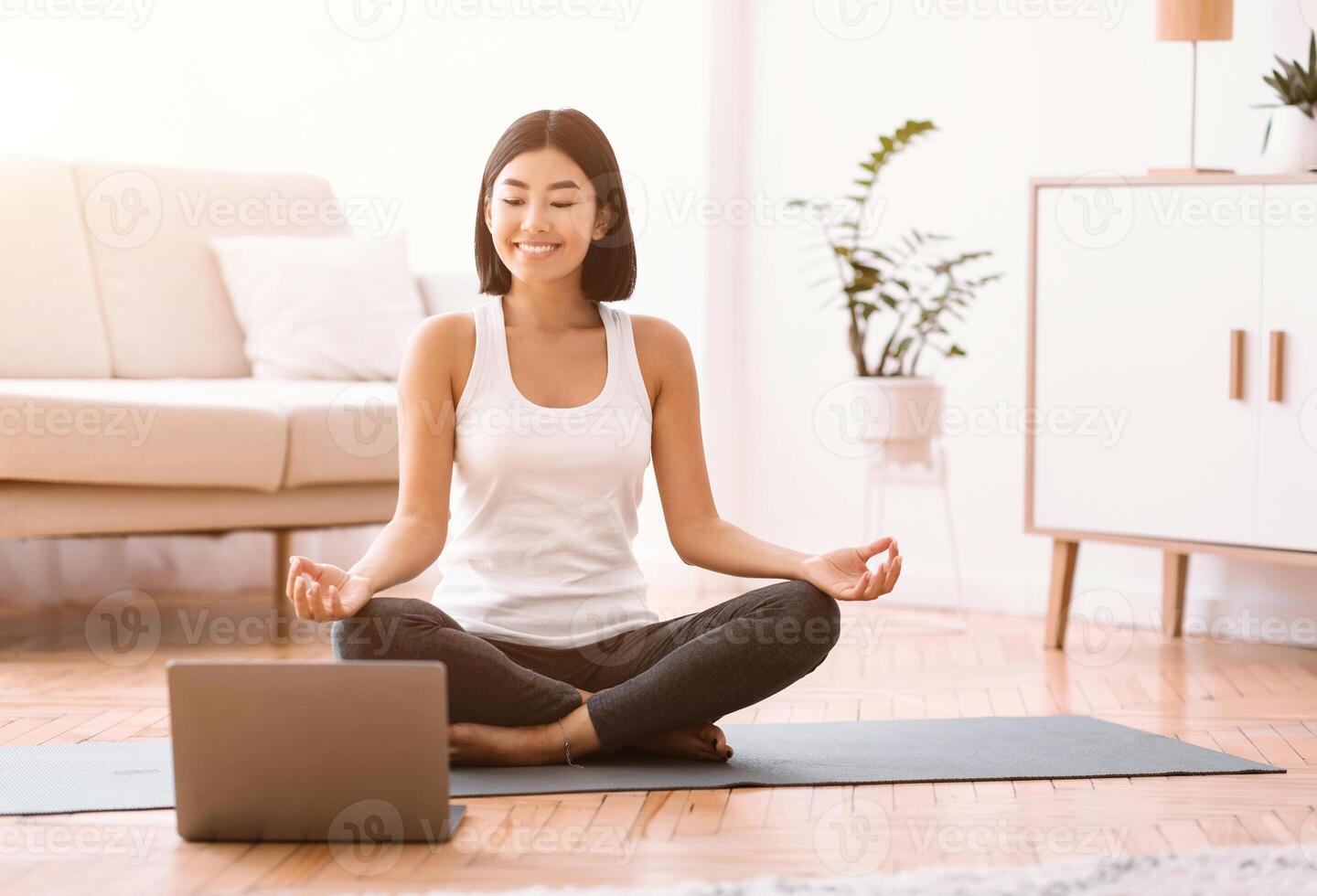 Keep Calm. Happy asian woman meditating with trainer online via laptop connection, copy space. Healthy lady sitting on the floor on yoga mat in lotus position, looking at computer and smiling photo