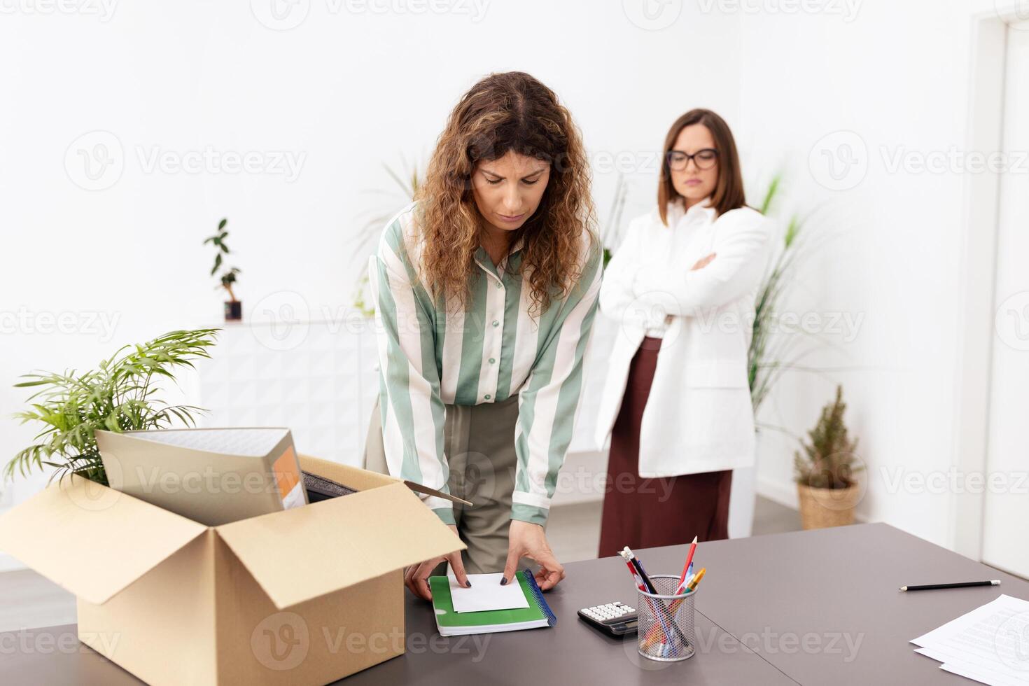 In a bright office space, two businesswomen are engaged in a conversation about work organization. One woman is packing items into a box while the other observes. photo