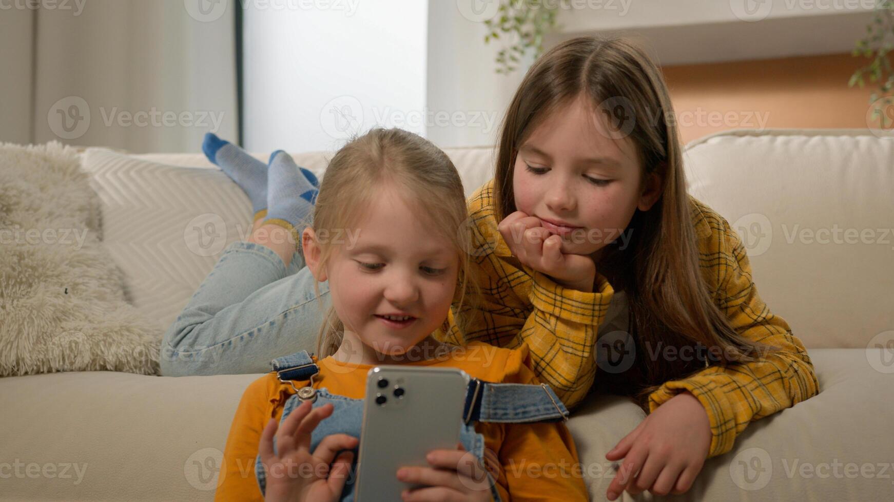 Two funny happy caucasian kids girls sisters friends children having fun playing on phone in living room couch play mobile online game together using smartphone browsing social network in Internet photo
