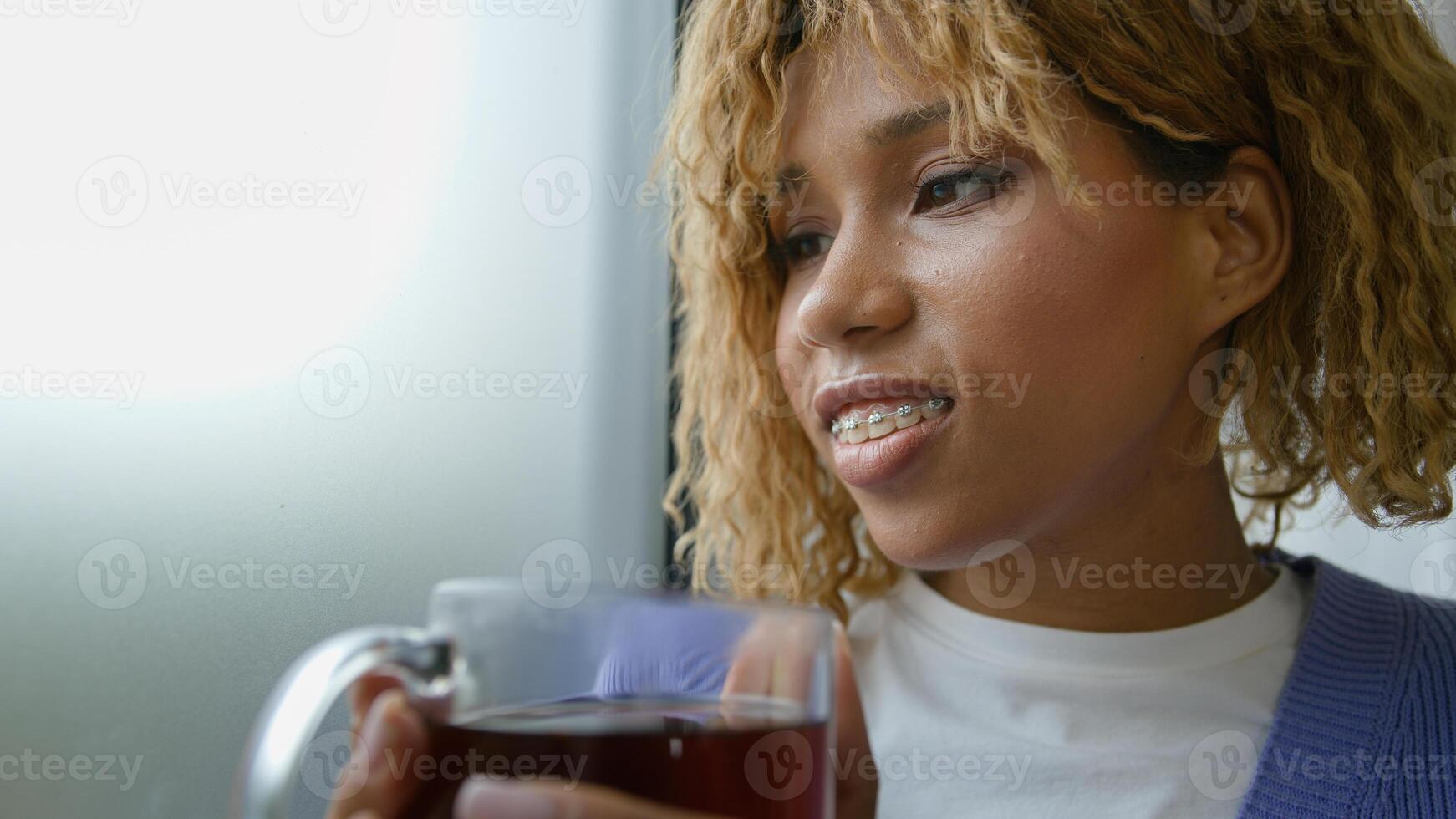 Happy smiling toothy with braces African American ethnic girl carefree lady joyful female woman at home looking at window relaxed harmony calm tranquil drinking tasty healthy herbal tea drink at home photo
