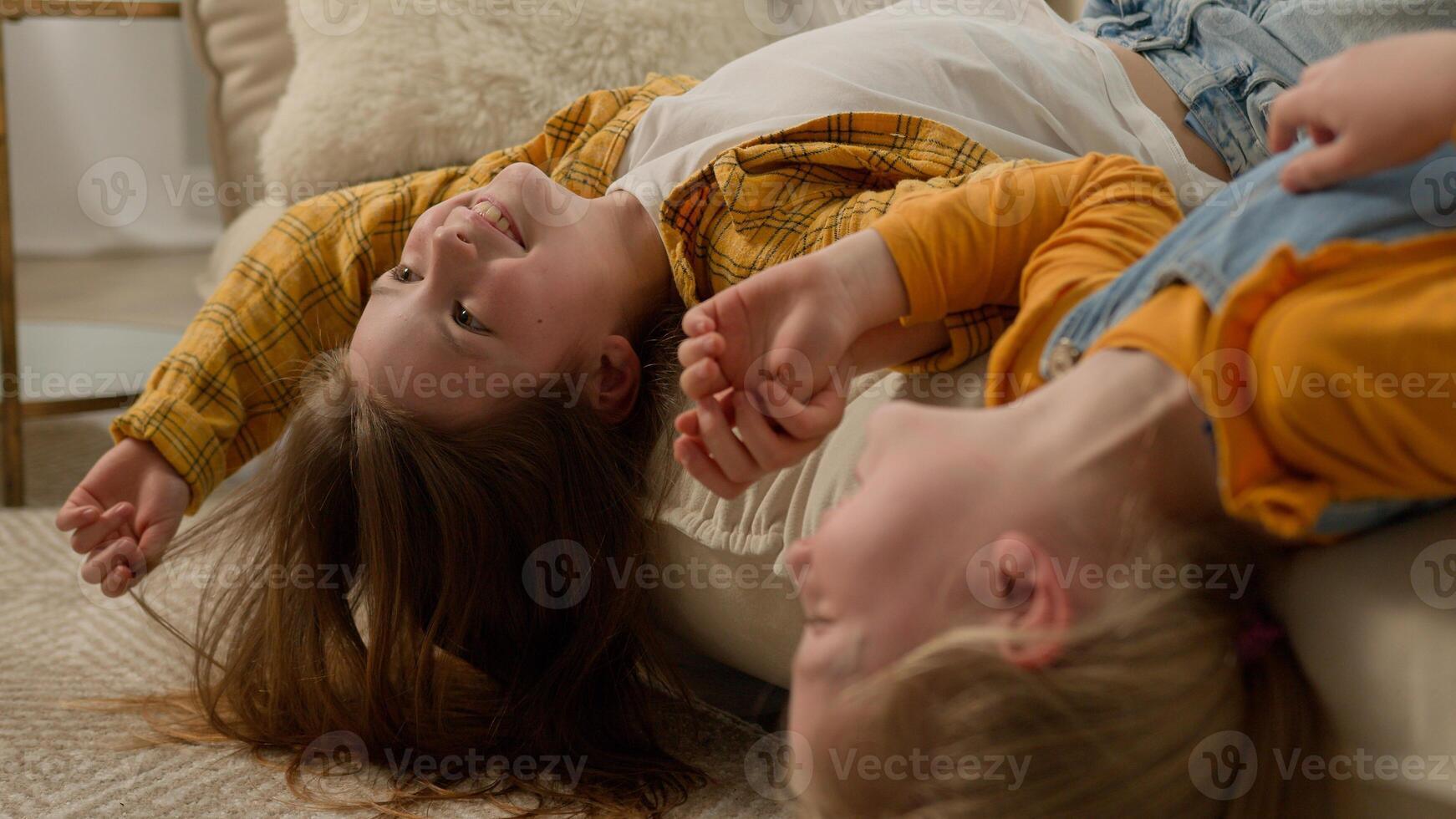 Smiling carefree Caucasian girls kids children sisters friends daughters lie upside down in living room couch playing fooling around together relax play laughing have fun weekend at home happy family photo