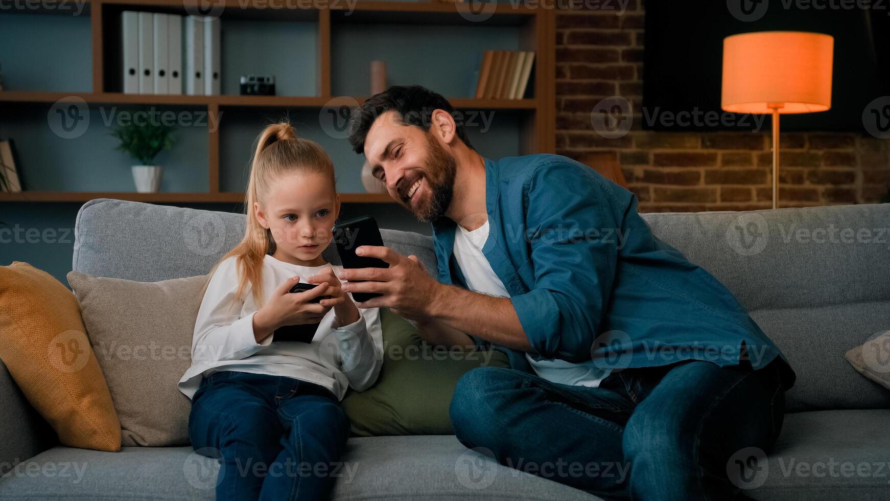 Caucasian man and little kid girl sit together at home hold modern phone browsing media content play games online adult dad teach child daughter use mobile apps showing funny on wireless device photo