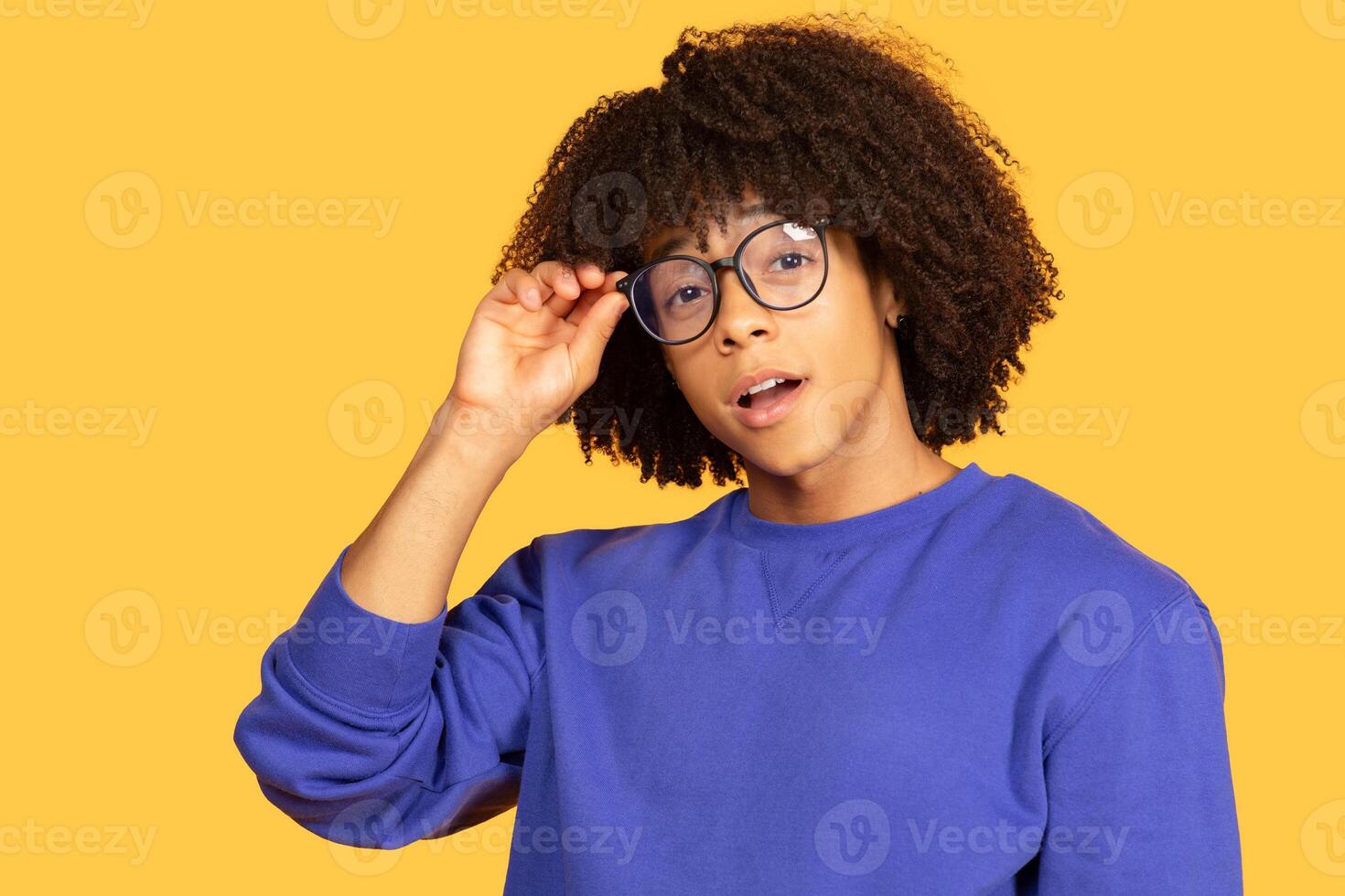 A young black man stands in front of a bright yellow backdrop. He sports curly hair and oversized glasses, conveying a trendy, playful vibe while casually adjusting his eyewear. photo