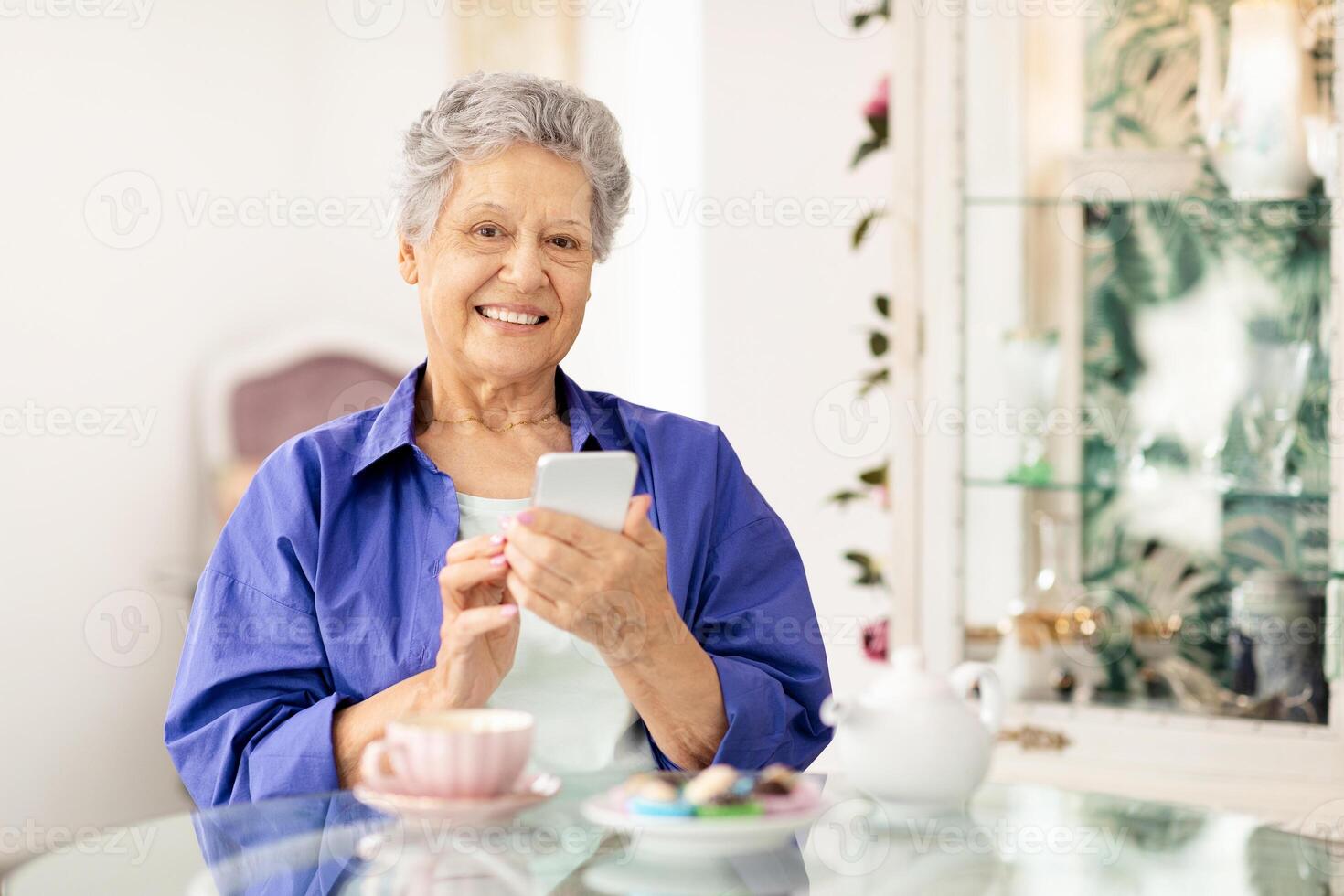 un mayor mujer con un calentar sonrisa se sienta a un vaso mesa en un brillante cafetería. ella disfruta un taza de café y usos su teléfono inteligente, rodeado por un atractivo atmósfera. foto